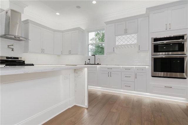 kitchen featuring wall chimney range hood, appliances with stainless steel finishes, ornamental molding, light hardwood / wood-style floors, and light stone counters