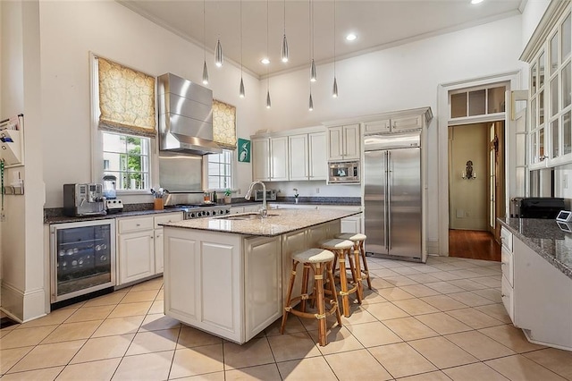 kitchen featuring wall chimney exhaust hood, built in appliances, a center island with sink, dark stone countertops, and beverage cooler