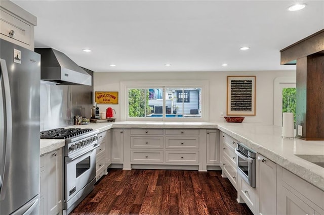 kitchen with wall chimney range hood, kitchen peninsula, appliances with stainless steel finishes, white cabinetry, and dark hardwood / wood-style floors