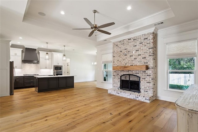 living room featuring a brick fireplace, ornamental molding, a raised ceiling, ceiling fan, and light hardwood / wood-style flooring