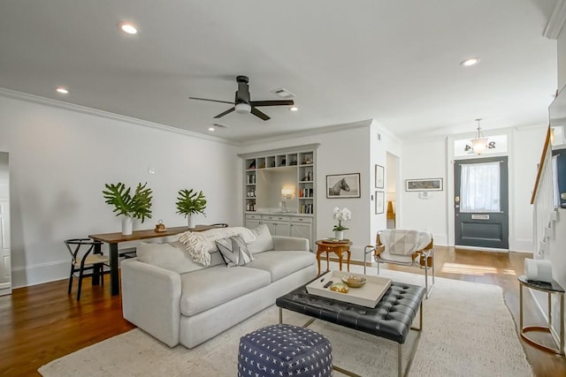 living room with built in shelves, wood-type flooring, ceiling fan, and crown molding