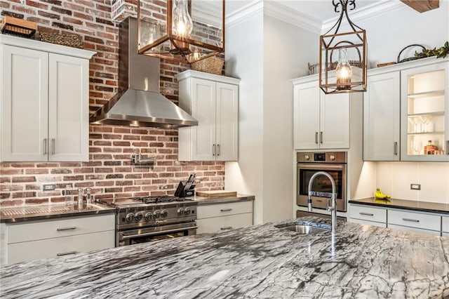 kitchen featuring hanging light fixtures, stainless steel stove, wall chimney range hood, white cabinetry, and dark stone countertops