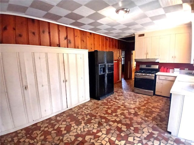 kitchen featuring black fridge with ice dispenser, wooden walls, white cabinetry, stainless steel gas range oven, and exhaust hood