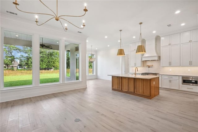 kitchen featuring white cabinets, an island with sink, pendant lighting, light hardwood / wood-style flooring, and premium range hood