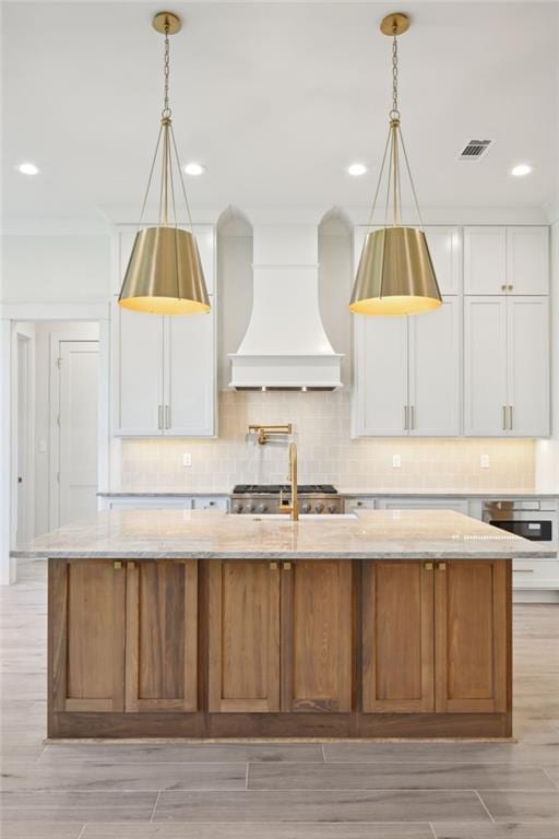 kitchen featuring white cabinets, custom range hood, a center island with sink, and light stone countertops