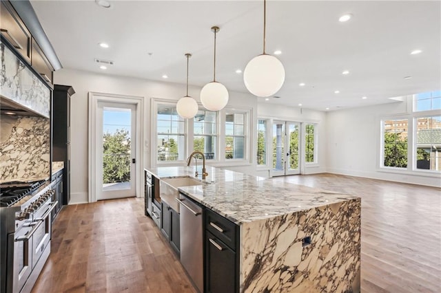 kitchen featuring sink, appliances with stainless steel finishes, hanging light fixtures, light stone countertops, and an island with sink