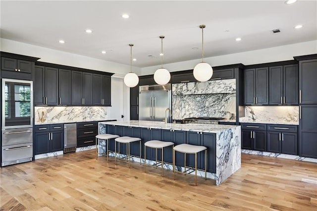 kitchen with stainless steel built in refrigerator, a kitchen bar, hanging light fixtures, an island with sink, and light stone countertops