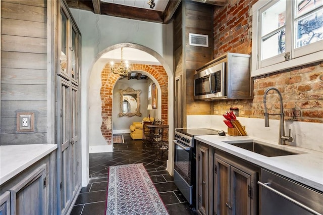 kitchen with stainless steel appliances, a notable chandelier, sink, beamed ceiling, and brick wall