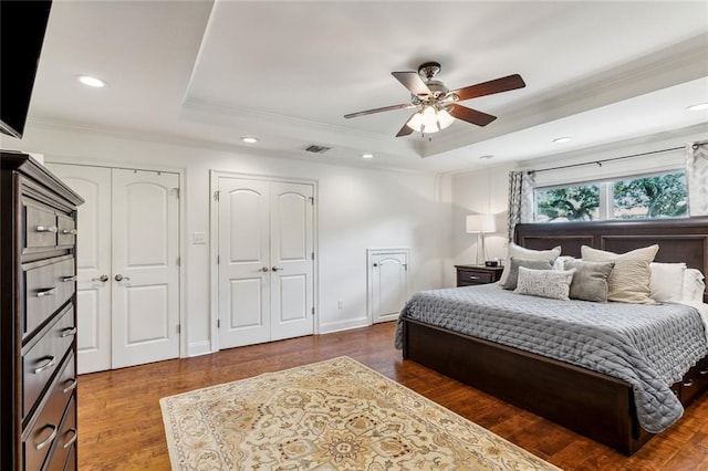 bedroom featuring ornamental molding, ceiling fan, a tray ceiling, dark wood-type flooring, and two closets