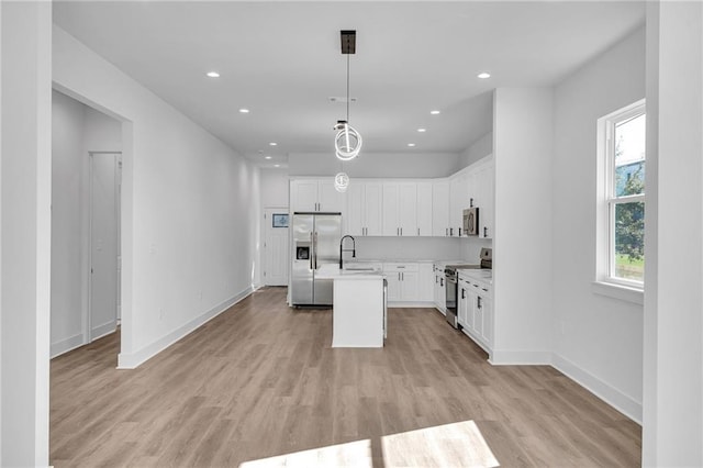 kitchen with appliances with stainless steel finishes, a kitchen island with sink, pendant lighting, light hardwood / wood-style floors, and white cabinetry