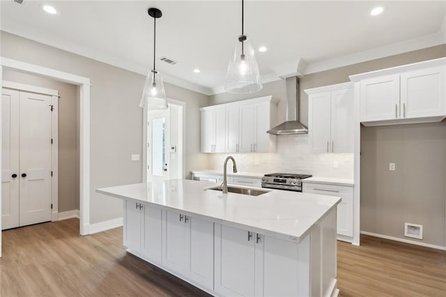 kitchen featuring a kitchen island with sink, sink, wall chimney range hood, pendant lighting, and white cabinets