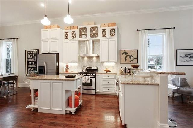 kitchen featuring appliances with stainless steel finishes, wall chimney exhaust hood, dark wood-type flooring, a center island, and hanging light fixtures