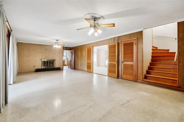 unfurnished living room with wood walls, a brick fireplace, ceiling fan, ornamental molding, and brick wall