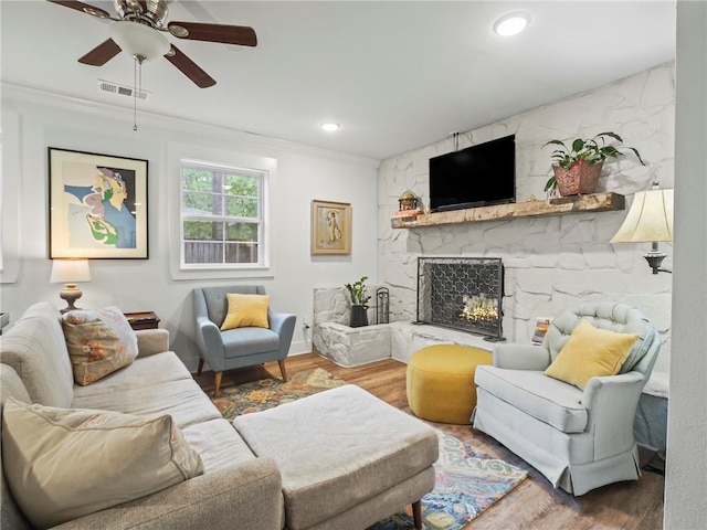 living room featuring a stone fireplace, crown molding, ceiling fan, and wood-type flooring