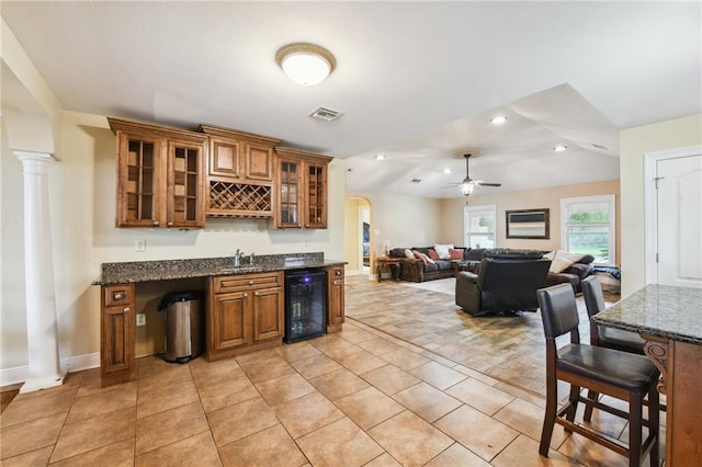 bar with wine cooler, ceiling fan, dark stone countertops, ornate columns, and light tile patterned floors