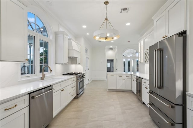 kitchen with high end appliances, premium range hood, white cabinets, sink, and decorative light fixtures