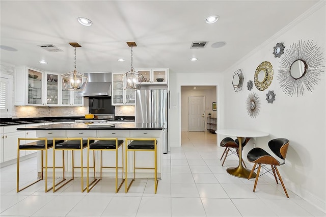 kitchen with crown molding, wall chimney range hood, white cabinets, stainless steel refrigerator, and hanging light fixtures