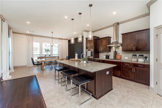 kitchen with pendant lighting, a kitchen island with sink, wall chimney range hood, crown molding, and light stone countertops