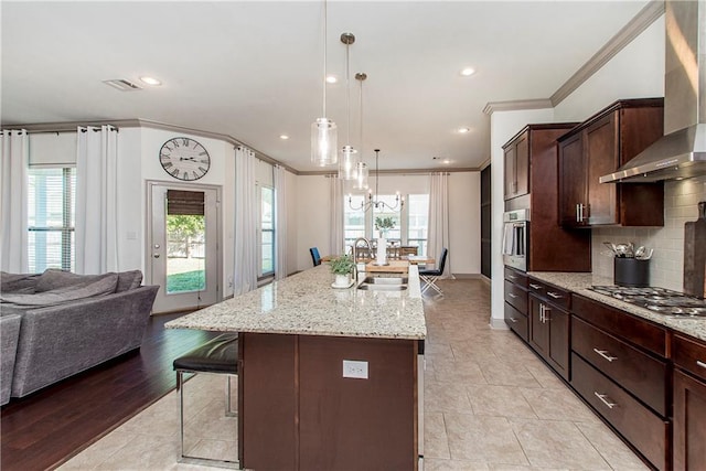 kitchen with sink, wall chimney range hood, light stone counters, an island with sink, and ornamental molding