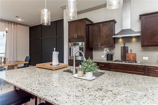 kitchen with wall chimney range hood, hanging light fixtures, ornamental molding, light stone counters, and stainless steel appliances