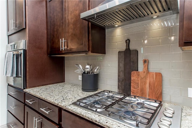 kitchen featuring light stone countertops, appliances with stainless steel finishes, tasteful backsplash, dark brown cabinets, and wall chimney range hood