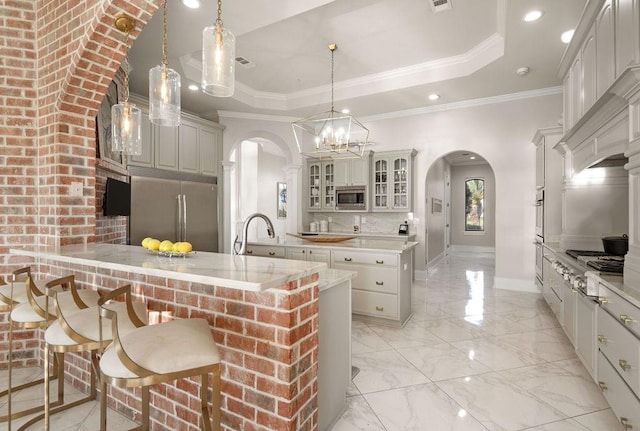 kitchen featuring pendant lighting, built in appliances, ornamental molding, an island with sink, and a tray ceiling