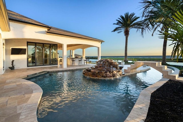 pool at dusk featuring a patio area, a water view, exterior bar, and exterior kitchen