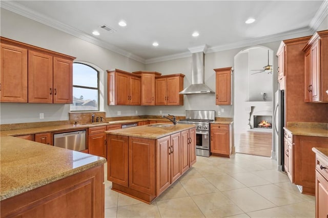 kitchen with appliances with stainless steel finishes, light stone counters, ornamental molding, wall chimney range hood, and an island with sink