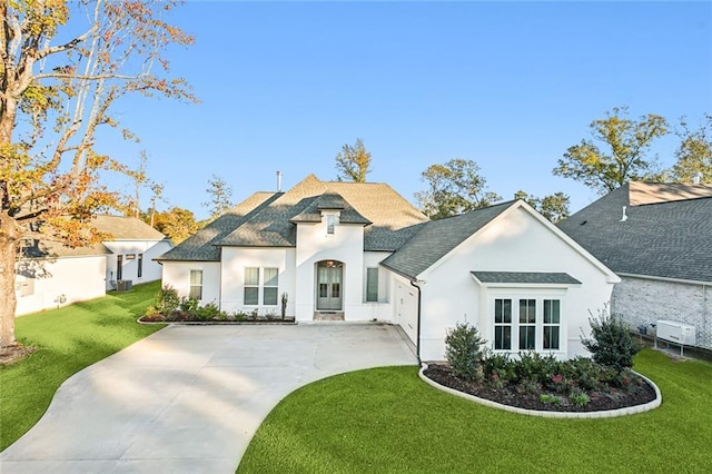 french country style house with a front yard, roof with shingles, driveway, and stucco siding