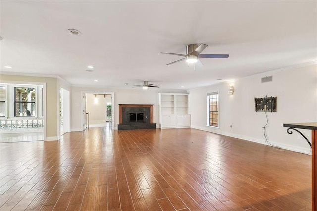 unfurnished living room featuring ceiling fan, built in features, wood-type flooring, and crown molding