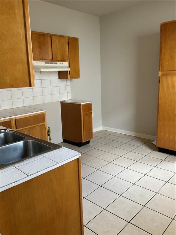 9009 Belfast St, New Orleans, LA 70118 - kitchen with under cabinet range hood, brown cabinetry, tile countertops, light tile patterned floors, and decorative backsplash
