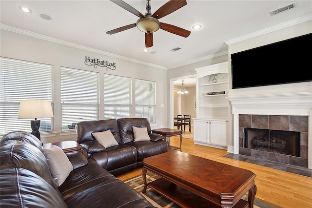 living room with a tile fireplace, ceiling fan with notable chandelier, crown molding, hardwood / wood-style flooring, and built in features