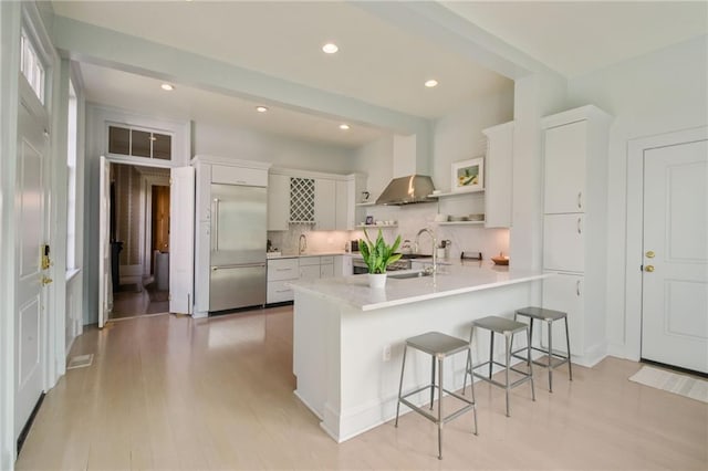 kitchen featuring built in fridge, white cabinets, wall chimney range hood, a kitchen breakfast bar, and kitchen peninsula