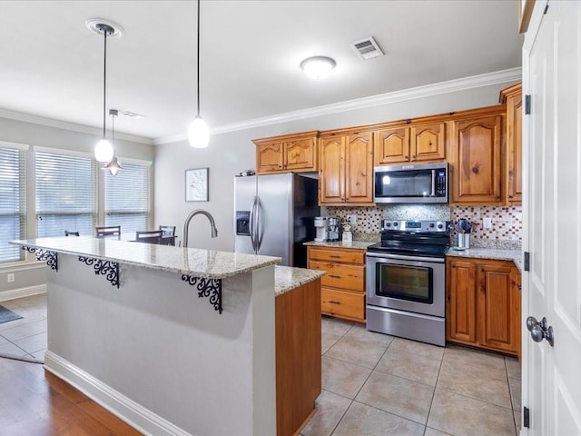 kitchen featuring a kitchen island with sink, hanging light fixtures, stainless steel appliances, and light stone counters