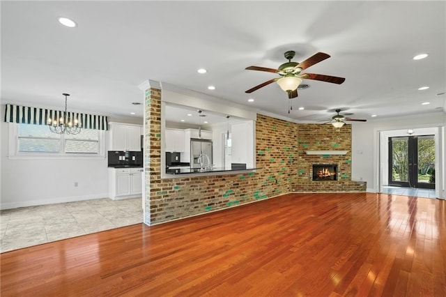 unfurnished living room with french doors, brick wall, a fireplace, ceiling fan with notable chandelier, and light wood-type flooring