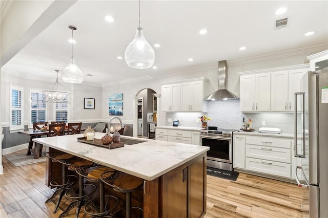 kitchen with sink, wall chimney range hood, pendant lighting, white cabinets, and appliances with stainless steel finishes