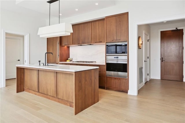 kitchen with sink, hanging light fixtures, an island with sink, appliances with stainless steel finishes, and light hardwood / wood-style floors