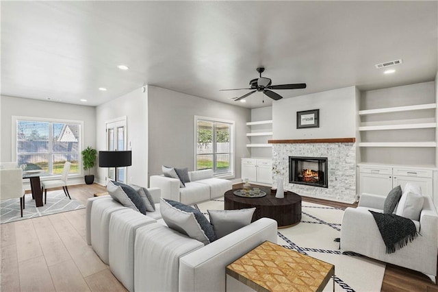 living room with built in features, light wood-type flooring, a stone fireplace, and ceiling fan