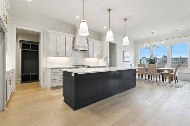 kitchen featuring pendant lighting, a kitchen island with sink, backsplash, ornamental molding, and white cabinets