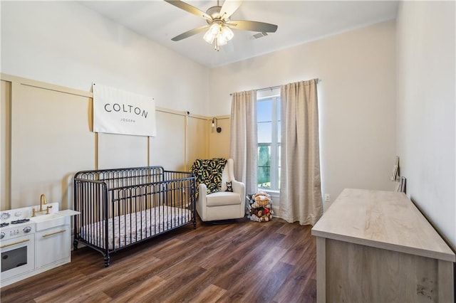 bedroom with a crib, ceiling fan, and dark wood-type flooring