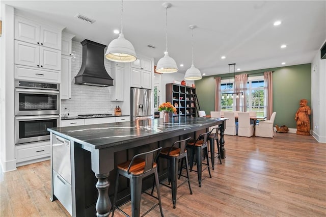 kitchen featuring a large island, appliances with stainless steel finishes, white cabinetry, custom range hood, and decorative light fixtures