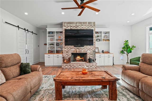 living room with built in shelves, a barn door, wood-type flooring, ceiling fan, and a fireplace