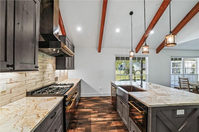 kitchen featuring sink, a center island with sink, appliances with stainless steel finishes, pendant lighting, and wall chimney range hood