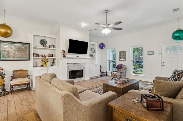 living room featuring a tile fireplace, ceiling fan, built in shelves, light hardwood / wood-style floors, and ornamental molding