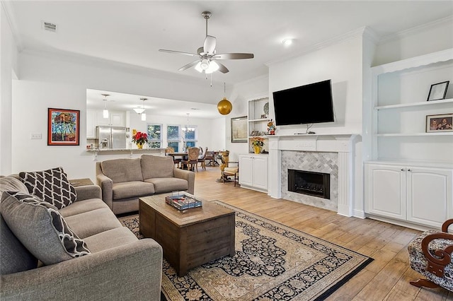 living room with a tile fireplace, ceiling fan, light hardwood / wood-style flooring, built in features, and crown molding