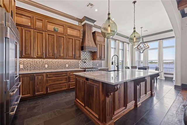 kitchen with premium range hood, sink, light stone counters, decorative light fixtures, and a kitchen island with sink