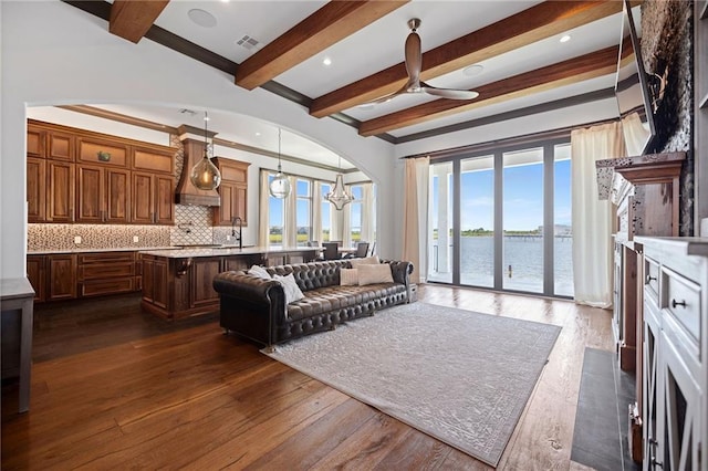 living room with sink, dark hardwood / wood-style floors, a water view, ceiling fan with notable chandelier, and beamed ceiling