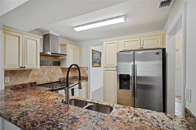 kitchen featuring appliances with stainless steel finishes, tasteful backsplash, wall chimney range hood, dark stone countertops, and cream cabinetry