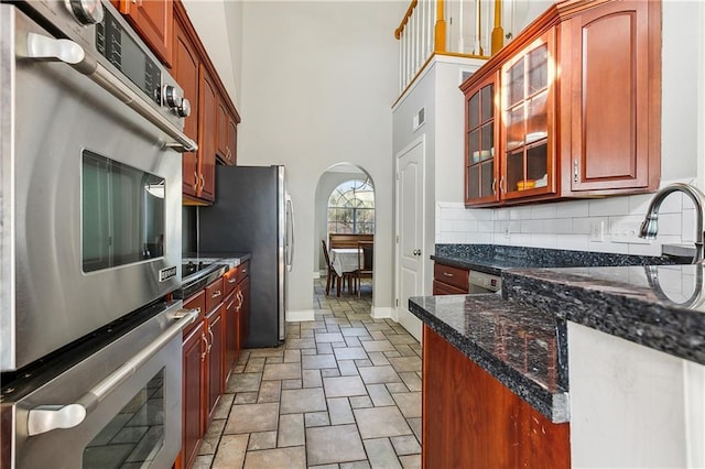 kitchen with a towering ceiling, backsplash, dark stone counters, stainless steel appliances, and sink