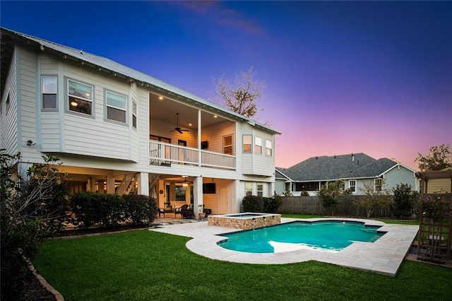 back house at dusk featuring ceiling fan, a balcony, a patio area, and a lawn